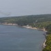 A military transport aircraft, appearing to be a C-130 Hercules, flies at low altitude over a tropical coastline. The coastline features dense palm tree vegetation, rocky cliffs, and a small coastal settlement visible along the shoreline. The image is taken from an elevated aerial perspective, possibly from another aircraft.