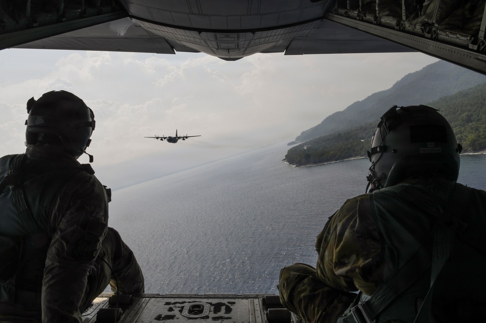 Two military personnel are seated at the open rear ramp of a military transport aircraft, observing another aircraft flying in formation ahead of them. The scene is captured from inside the cargo bay looking outward, with a coastal landscape featuring green hills and open water visible in the background. Both crew members are wearing flight helmets and military gear.