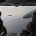 Two military personnel are seated at the open rear ramp of a military transport aircraft, observing another aircraft flying in formation ahead of them. The scene is captured from inside the cargo bay looking outward, with a coastal landscape featuring green hills and open water visible in the background. Both crew members are wearing flight helmets and military gear.
