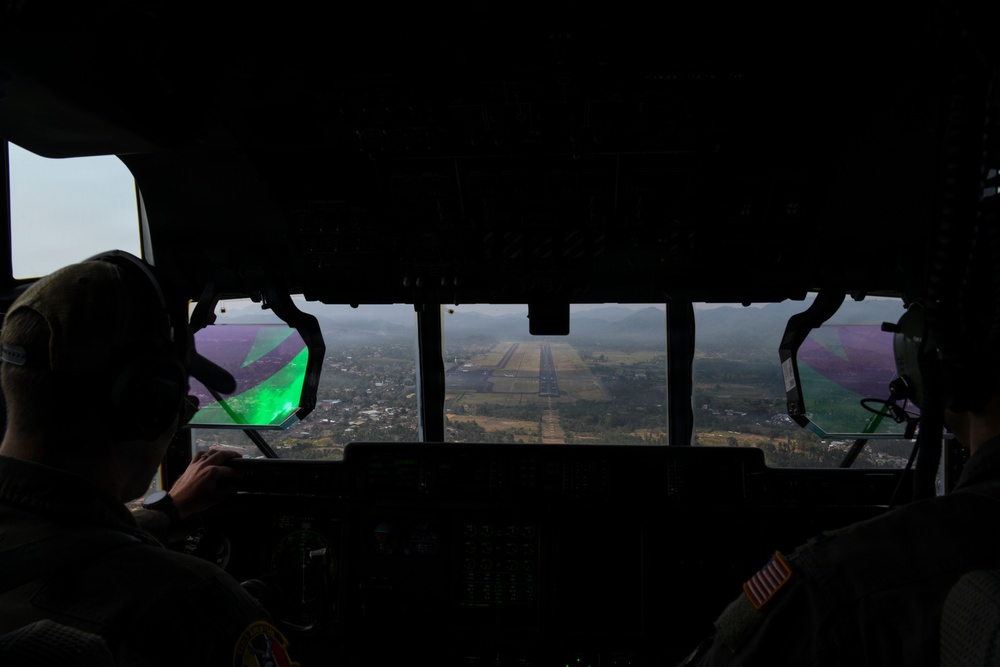 View from inside a military aircraft cockpit on final approach to a runway. Two crew members are visible in silhouette, with one on the left wearing a headset and the other on the right displaying a US flag patch on their flight suit. A green laser or optical reflection is visible on the left side of the windscreen, which may indicate a laser strike directed at the aircraft.