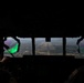 View from inside a military aircraft cockpit on final approach to a runway. Two crew members are visible in silhouette, with one on the left wearing a headset and the other on the right displaying a US flag patch on their flight suit. A green laser or optical reflection is visible on the left side of the windscreen, which may indicate a laser strike directed at the aircraft.