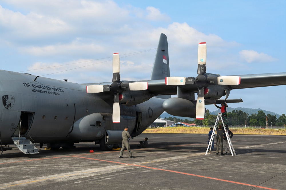 A gray military transport aircraft belonging to the Indonesian Air Force (TNI Angkatan Udara) is parked on a tarmac, undergoing maintenance. Ground crew personnel are working around the aircraft, including one technician on a ladder inspecting or servicing one of the rear propeller engines. The aircraft displays Indonesian Air Force markings and the number '33' on its fuselage.