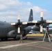A gray military transport aircraft belonging to the Indonesian Air Force (TNI Angkatan Udara) is parked on a tarmac, undergoing maintenance. Ground crew personnel are working around the aircraft, including one technician on a ladder inspecting or servicing one of the rear propeller engines. The aircraft displays Indonesian Air Force markings and the number '33' on its fuselage.
