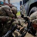 Military personnel in MultiCam/OCP camouflage uniforms are conducting what appears to be a tactical combat casualty care (TCCC) training exercise. One soldier is lying on the ground simulating a casualty while two others provide simulated medical treatment. The scene takes place near a military HMMWV (Humvee) vehicle, with additional personnel observing in the background.