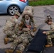 Three soldiers in OCP (Operational Camouflage Pattern) uniforms are performing a tactical casualty care drill on a fourth soldier lying on the pavement. Two soldiers are attending to the simulated casualty while a third prepares a folding litter/stretcher for transport. This appears to be a combat lifesaver or tactical combat casualty care (TCCC) training exercise conducted in a parking lot environment.