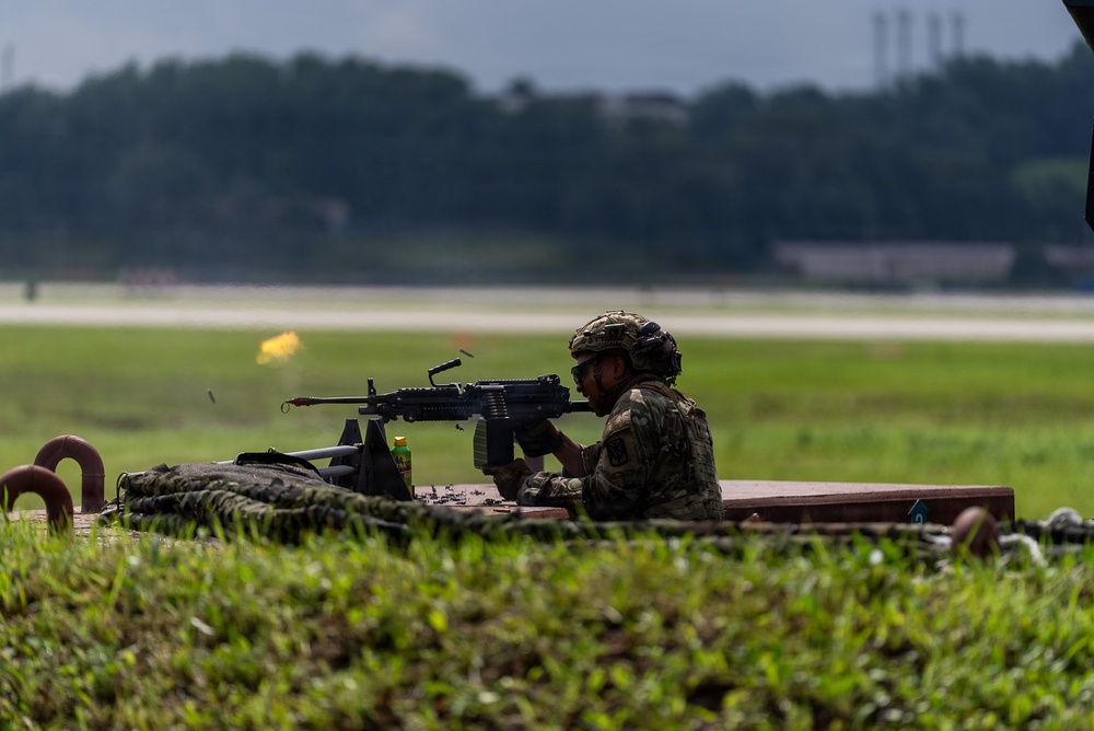 A soldier in full combat gear is firing a machine gun from a prone or low-profile position on what appears to be a raised platform or berm. A muzzle flash is visible at the barrel, indicating active firing. The setting appears to be a military firing range adjacent to an airfield or runway.