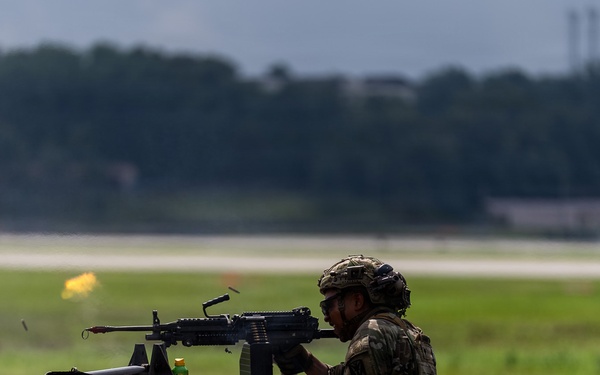 A soldier in full combat gear is firing a machine gun from a prone or low-profile position on what appears to be a raised platform or berm. A muzzle flash is visible at the barrel, indicating active firing. The setting appears to be a military firing range adjacent to an airfield or runway.
