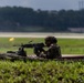 A soldier in full combat gear is firing a machine gun from a prone or low-profile position on what appears to be a raised platform or berm. A muzzle flash is visible at the barrel, indicating active firing. The setting appears to be a military firing range adjacent to an airfield or runway.