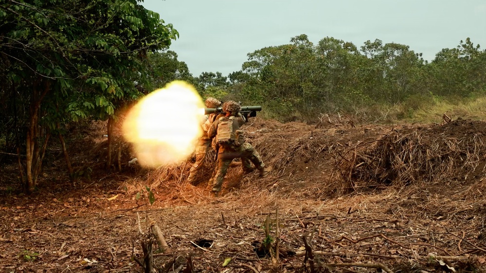 Two soldiers in camouflage uniforms and tactical vests are firing a shoulder-launched rocket or missile system, producing a large muzzle blast fireball to the left. The scene takes place in a cleared dirt area at the edge of a tropical or subtropical woodland. This appears to be a military training exercise based on the controlled environment and positioning.