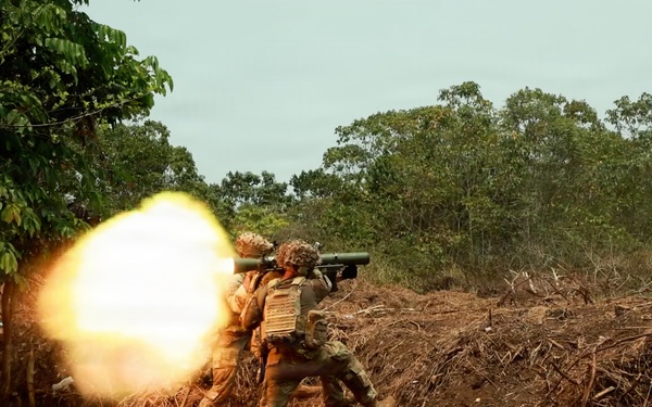 Two soldiers in camouflage uniforms and tactical vests are firing a shoulder-launched rocket or missile system, producing a large muzzle blast fireball to the left. The scene takes place in a cleared dirt area at the edge of a tropical or subtropical woodland. This appears to be a military training exercise based on the controlled environment and positioning.