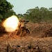 Two soldiers in camouflage uniforms and tactical vests are firing a shoulder-launched rocket or missile system, producing a large muzzle blast fireball to the left. The scene takes place in a cleared dirt area at the edge of a tropical or subtropical woodland. This appears to be a military training exercise based on the controlled environment and positioning.