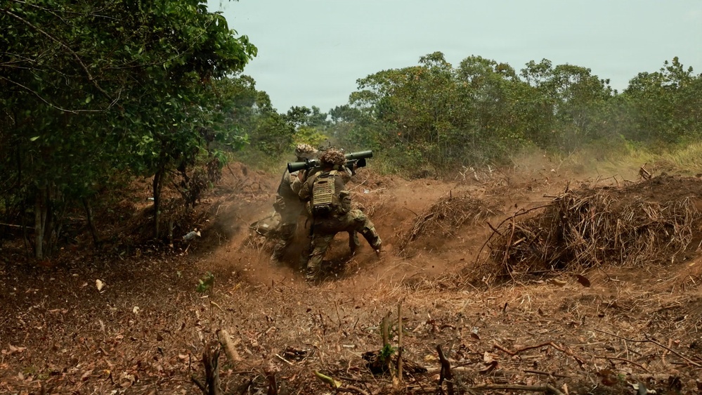 Two soldiers in camouflage uniforms are operating in a cleared dirt area surrounded by tropical vegetation. One soldier is shouldering what appears to be a rocket launcher or recoilless rifle, while dust and debris are visible around them, suggesting recent or imminent firing. The setting appears to be a military training exercise in a tropical or subtropical environment.