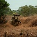 Two soldiers in camouflage uniforms are operating in a cleared dirt area surrounded by tropical vegetation. One soldier is shouldering what appears to be a rocket launcher or recoilless rifle, while dust and debris are visible around them, suggesting recent or imminent firing. The setting appears to be a military training exercise in a tropical or subtropical environment.