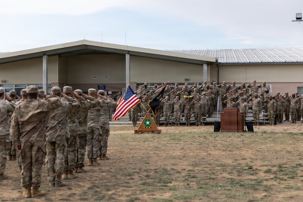 A formal U.S. Army military ceremony is taking place outdoors on a parade field in front of a military installation building. Soldiers in Army Combat Uniforms (ACUs) are rendered in a salute formation, with color guard flags including the U.S. national flag and a unit guidon displayed on a ceremonial stand. The event appears to be a change of command, activation, or similar official military ceremony.
