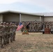 A formal U.S. Army military ceremony is taking place outdoors on a parade field in front of a military installation building. Soldiers in Army Combat Uniforms (ACUs) are rendered in a salute formation, with color guard flags including the U.S. national flag and a unit guidon displayed on a ceremonial stand. The event appears to be a change of command, activation, or similar official military ceremony.