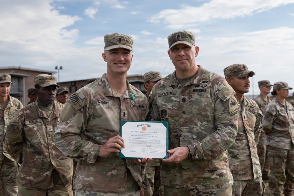 Two U.S. Army soldiers stand together holding a Department of the Army Army Commendation Medal certificate during what appears to be a formal awards ceremony. The soldier on the left, identified by nametape as 'ASBURY,' is wearing a commendation medal ribbon on his uniform. They are surrounded by other uniformed soldiers standing in formation in an outdoor military installation.