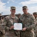 Two U.S. Army soldiers stand together holding a Department of the Army Army Commendation Medal certificate during what appears to be a formal awards ceremony. The soldier on the left, identified by nametape as 'ASBURY,' is wearing a commendation medal ribbon on his uniform. They are surrounded by other uniformed soldiers standing in formation in an outdoor military installation.