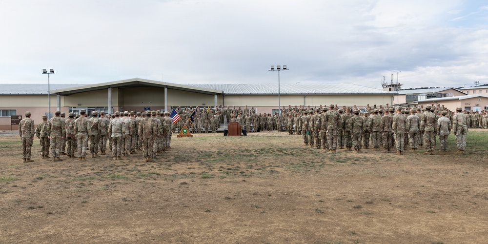 A large military formation ceremony is taking place on an outdoor parade ground at what appears to be a U.S. Army installation. Hundreds of soldiers in Army Combat Uniforms (ACUs/OCP pattern) are arranged in formation facing a central podium, with the American flag and unit colors visible. The setting includes low-rise military buildings in the background under an overcast sky.