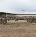 A large military formation ceremony is taking place on an outdoor parade ground at what appears to be a U.S. Army installation. Hundreds of soldiers in Army Combat Uniforms (ACUs/OCP pattern) are arranged in formation facing a central podium, with the American flag and unit colors visible. The setting includes low-rise military buildings in the background under an overcast sky.