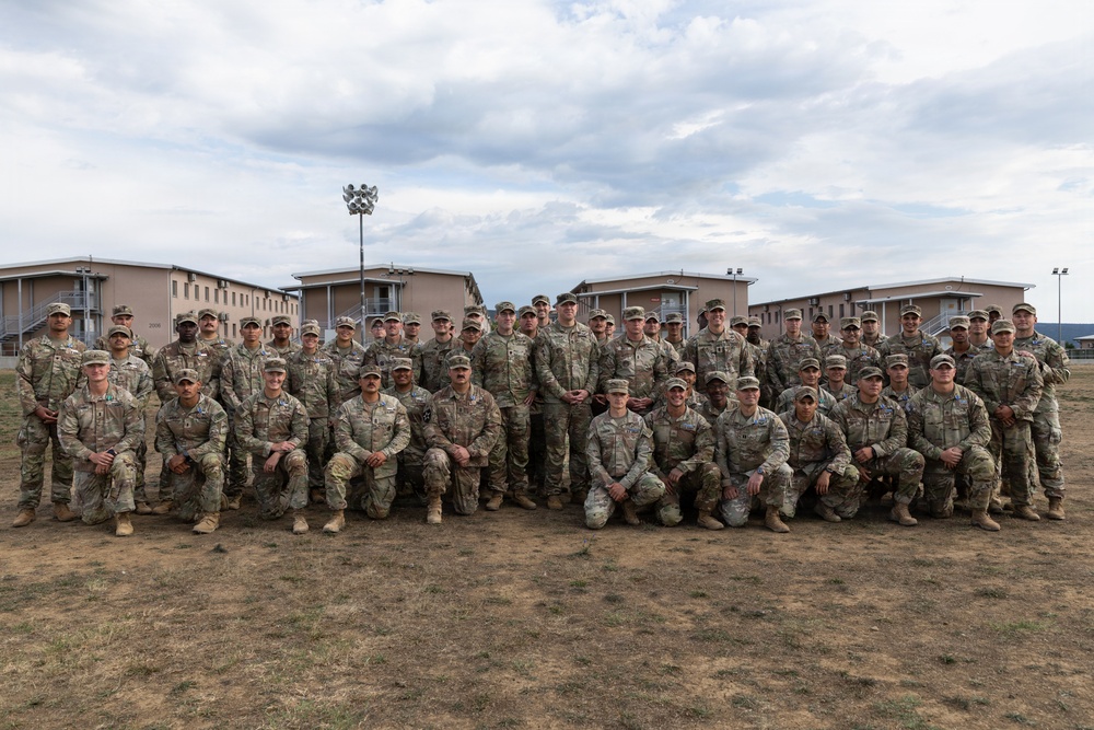 A large group of approximately 40-50 military personnel in U.S. Army Operational Camouflage Pattern (OCP) uniforms pose for a formal group photograph outdoors. They are arranged in two rows — one standing and one kneeling — on a dry grass field in front of what appear to be military barracks or administrative buildings. The overcast sky and arid terrain suggest a military installation, possibly in a semi-arid region.