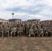 A large group of approximately 40-50 military personnel in U.S. Army Operational Camouflage Pattern (OCP) uniforms pose for a formal group photograph outdoors. They are arranged in two rows — one standing and one kneeling — on a dry grass field in front of what appear to be military barracks or administrative buildings. The overcast sky and arid terrain suggest a military installation, possibly in a semi-arid region.