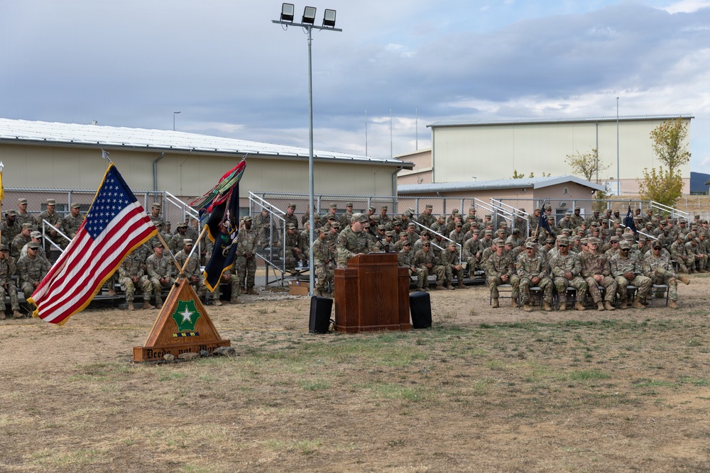 A formal U.S. Army military ceremony is taking place outdoors on a base installation. A soldier is speaking at a wooden podium while a large formation of soldiers in Army Combat Uniforms (ACUs) are seated on bleachers and chairs. The American flag and unit colors are displayed on a triangular wooden flag stand in the foreground.