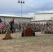 A formal U.S. Army military ceremony is taking place outdoors on a base installation. A soldier is speaking at a wooden podium while a large formation of soldiers in Army Combat Uniforms (ACUs) are seated on bleachers and chairs. The American flag and unit colors are displayed on a triangular wooden flag stand in the foreground.