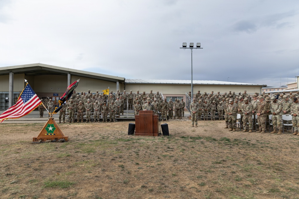 A military ceremony is taking place outdoors on a military installation, with a large formation of soldiers in Army Combat Uniforms (ACU/OCP pattern) assembled in front of a building. A speaker stands at a wooden podium while flags, including the U.S. flag and unit colors, are displayed on a stand to the left. The wooden base of the flag stand bears an insignia and text, suggesting a unit-specific ceremony such as a change of command or activation/inactivation.