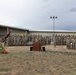 A military ceremony is taking place outdoors on a military installation, with a large formation of soldiers in Army Combat Uniforms (ACU/OCP pattern) assembled in front of a building. A speaker stands at a wooden podium while flags, including the U.S. flag and unit colors, are displayed on a stand to the left. The wooden base of the flag stand bears an insignia and text, suggesting a unit-specific ceremony such as a change of command or activation/inactivation.