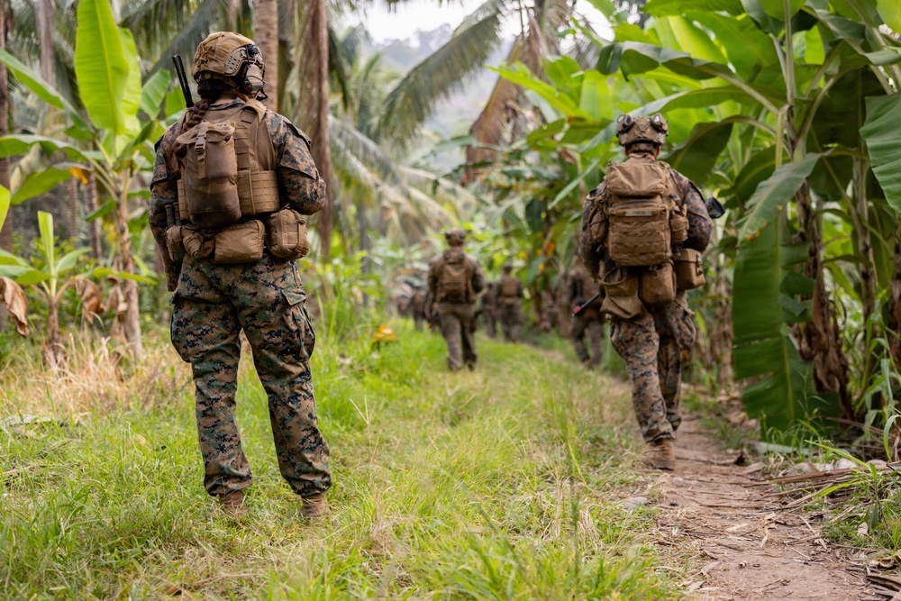 A patrol of military personnel in full combat gear moves along a narrow dirt trail through dense tropical vegetation, including banana trees and palm trees. The soldiers are wearing matching digital camouflage uniforms consistent with U.S. Marine Corps MARPAT pattern, carrying tactical backpacks and equipment. The formation suggests a structured patrol movement through a jungle or tropical rural environment.