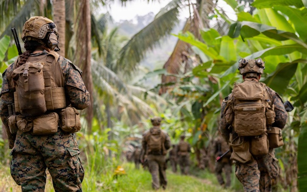 A patrol of military personnel in full combat gear moves along a narrow dirt trail through dense tropical vegetation, including banana trees and palm trees. The soldiers are wearing matching digital camouflage uniforms consistent with U.S. Marine Corps MARPAT pattern, carrying tactical backpacks and equipment. The formation suggests a structured patrol movement through a jungle or tropical rural environment.