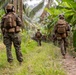 A patrol of military personnel in full combat gear moves along a narrow dirt trail through dense tropical vegetation, including banana trees and palm trees. The soldiers are wearing matching digital camouflage uniforms consistent with U.S. Marine Corps MARPAT pattern, carrying tactical backpacks and equipment. The formation suggests a structured patrol movement through a jungle or tropical rural environment.