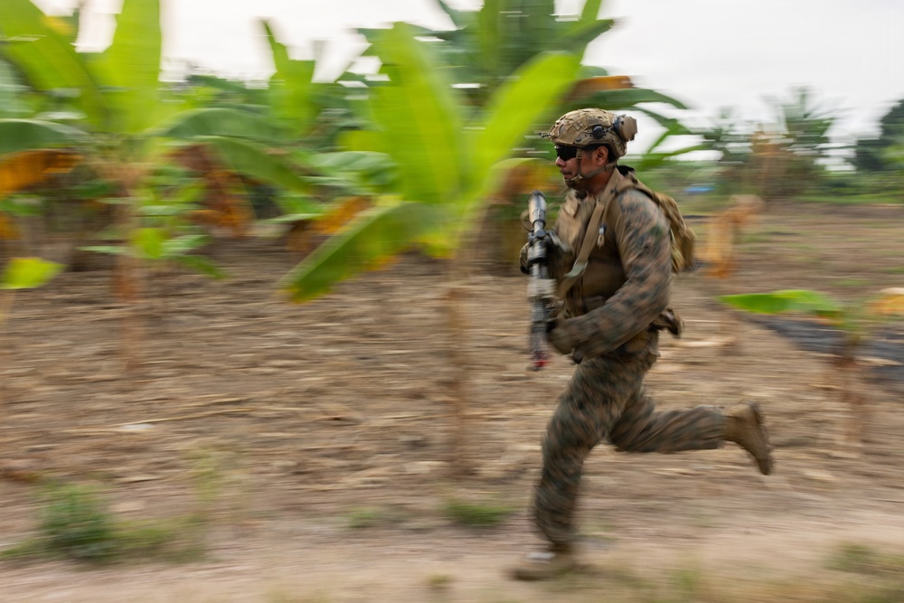 A soldier in full combat gear is captured mid-sprint across an open dirt area surrounded by tropical vegetation, likely banana plants. The image uses a panning photography technique, keeping the subject relatively sharp while blurring the background to convey speed and motion. The setting appears to be a tropical or subtropical environment during a military exercise or operation.