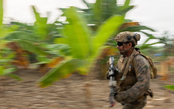 A soldier in full combat gear is captured mid-sprint across an open dirt area surrounded by tropical vegetation, likely banana plants. The image uses a panning photography technique, keeping the subject relatively sharp while blurring the background to convey speed and motion. The setting appears to be a tropical or subtropical environment during a military exercise or operation.