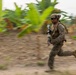 A soldier in full combat gear is captured mid-sprint across an open dirt area surrounded by tropical vegetation, likely banana plants. The image uses a panning photography technique, keeping the subject relatively sharp while blurring the background to convey speed and motion. The setting appears to be a tropical or subtropical environment during a military exercise or operation.