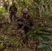 Three military personnel in full combat gear are conducting a patrol through dense jungle terrain. The lead soldier is in a crouched, tactical position holding an assault rifle with an optic scope, while two others follow behind in a column formation. The environment is a tropical forest with heavy undergrowth, fallen leaves, and dense vegetation.