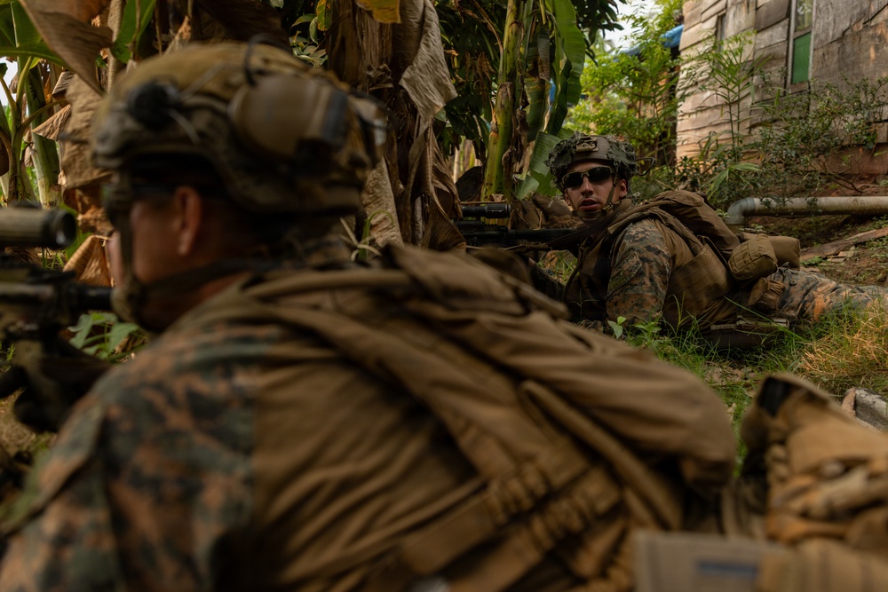 Two military personnel in MARPAT (Marine pattern) camouflage uniforms are positioned in a low prone or crouched stance among vegetation near a structure. The foreground soldier is equipped with a scoped rifle, while the background soldier appears to be aiming a weapon and wearing sunglasses. The setting suggests a training exercise or tactical operation in a semi-urban environment with tropical vegetation.
