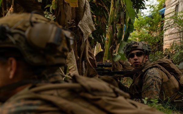Two military personnel in MARPAT (Marine pattern) camouflage uniforms are positioned in a low prone or crouched stance among vegetation near a structure. The foreground soldier is equipped with a scoped rifle, while the background soldier appears to be aiming a weapon and wearing sunglasses. The setting suggests a training exercise or tactical operation in a semi-urban environment with tropical vegetation.