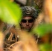A military service member is photographed through dense jungle foliage, partially concealed by vegetation. The soldier is wearing tactical gear including a helmet with camouflage netting and dark ballistic sunglasses. The image appears to capture a reconnaissance or concealment exercise in a tropical environment.