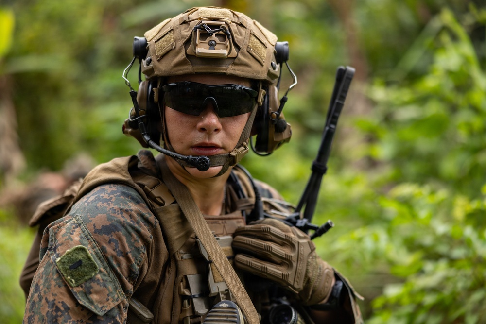 A military service member in full combat gear is photographed in a jungle or forested environment. The individual is wearing USMC MARPAT woodland camouflage, a tan tactical helmet with mounted accessories, and tactical hearing protection with a boom microphone. The image is a close-up portrait-style shot emphasizing the soldier's equipment and alert posture.