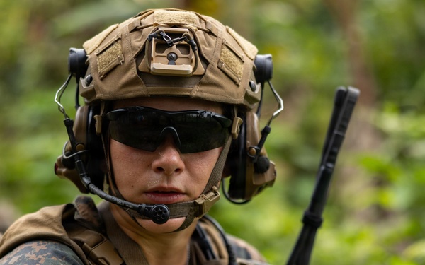 A military service member in full combat gear is photographed in a jungle or forested environment. The individual is wearing USMC MARPAT woodland camouflage, a tan tactical helmet with mounted accessories, and tactical hearing protection with a boom microphone. The image is a close-up portrait-style shot emphasizing the soldier's equipment and alert posture.