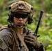 A military service member in full combat gear is photographed in a jungle or forested environment. The individual is wearing USMC MARPAT woodland camouflage, a tan tactical helmet with mounted accessories, and tactical hearing protection with a boom microphone. The image is a close-up portrait-style shot emphasizing the soldier's equipment and alert posture.