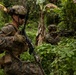Two military personnel in MARPAT (Marine pattern) camouflage uniforms are conducting a patrol or tactical exercise in a dense jungle/tropical environment. The foreground soldier is in a kneeling security position while the background soldier is aiming their rifle. The lush green vegetation and banana trees suggest a tropical location, consistent with a training exercise or operational deployment.