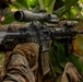 A military service member in MARPAT (Marine pattern) camouflage uniform is aiming an AR-platform rifle equipped with a magnified optic scope and vertical foregrip in a jungle or tropical environment. The weapon appears to be an M4/M16-variant with multiple rail-mounted accessories. The setting suggests a training exercise or operational deployment in a tropical region.