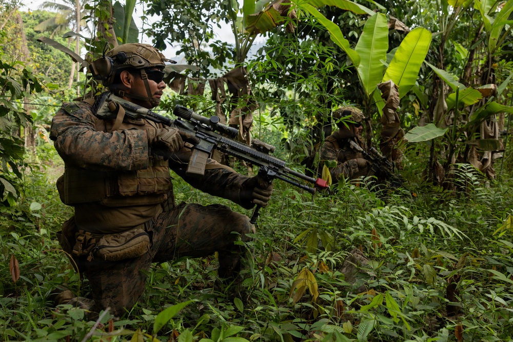 Two military personnel in MARPAT (Marine pattern) camouflage uniforms are conducting what appears to be a jungle patrol or training exercise in a dense tropical environment with banana trees and heavy vegetation. The foreground soldier is in a kneeling position aiming a rifle equipped with an optic scope, while a second soldier is visible in the background in a standing position. The setting and uniforms suggest a U.S. Marine Corps exercise in a tropical region.