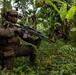 Two military personnel in MARPAT (Marine pattern) camouflage uniforms are conducting what appears to be a jungle patrol or training exercise in a dense tropical environment with banana trees and heavy vegetation. The foreground soldier is in a kneeling position aiming a rifle equipped with an optic scope, while a second soldier is visible in the background in a standing position. The setting and uniforms suggest a U.S. Marine Corps exercise in a tropical region.