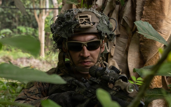 A soldier in full combat gear is positioned in dense jungle vegetation, holding a rifle in a ready stance. The individual is wearing MARPAT (Marine pattern) camouflage uniform, a tactical helmet with foliage camouflage cover and mounted equipment, and dark sunglasses. The setting appears to be a tropical jungle environment, consistent with a military training exercise or patrol operation.