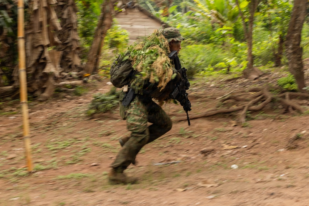 A soldier in full camouflage gear, including a ghillie suit with natural vegetation attached to their pack, is running at speed through a jungle or tropical environment. The motion blur effect indicates rapid movement, captured with a panning camera technique. A rustic wooden structure and dense tropical vegetation are visible in the background.