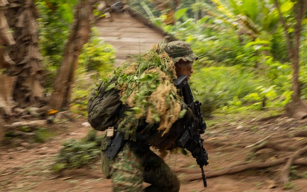 A soldier in full camouflage gear, including a ghillie suit with natural vegetation attached to their pack, is running at speed through a jungle or tropical environment. The motion blur effect indicates rapid movement, captured with a panning camera technique. A rustic wooden structure and dense tropical vegetation are visible in the background.