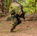 A soldier in full camouflage gear, including a ghillie suit with natural vegetation attached to their pack, is running at speed through a jungle or tropical environment. The motion blur effect indicates rapid movement, captured with a panning camera technique. A rustic wooden structure and dense tropical vegetation are visible in the background.